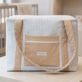 A soft fabric tote bag with a light blue and white vertical stripe pattern, tan side panels and handles, and a tan front zip pocket with a small white brand label, displayed on a light wood pedestal in a softly blurred nursery-like setting.