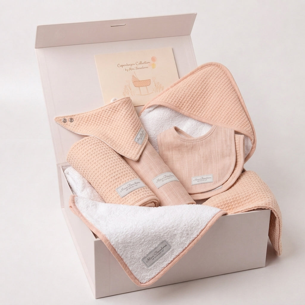 A baby gift set in a light pink box, containing peach-colored waffle-weave cloths and a white terry cloth, with a brand card inside the box.