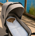 A close-up of a stroller bassinet setup outdoors by a lake, featuring a beige stroller body with a light blue and white striped mattress and a tan hood insert.