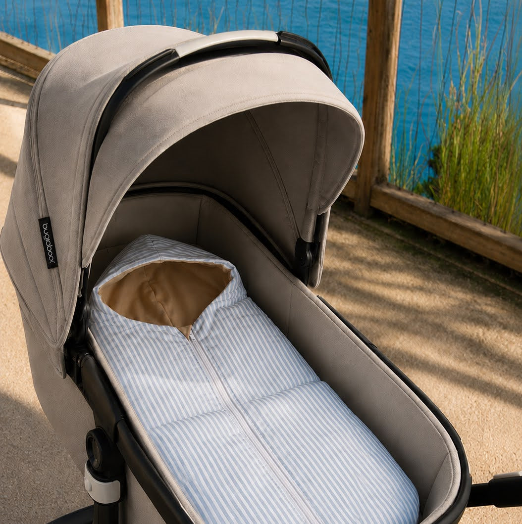 A close-up of a stroller bassinet setup outdoors by a lake, featuring a beige stroller body with a light blue and white striped mattress and a tan hood insert.