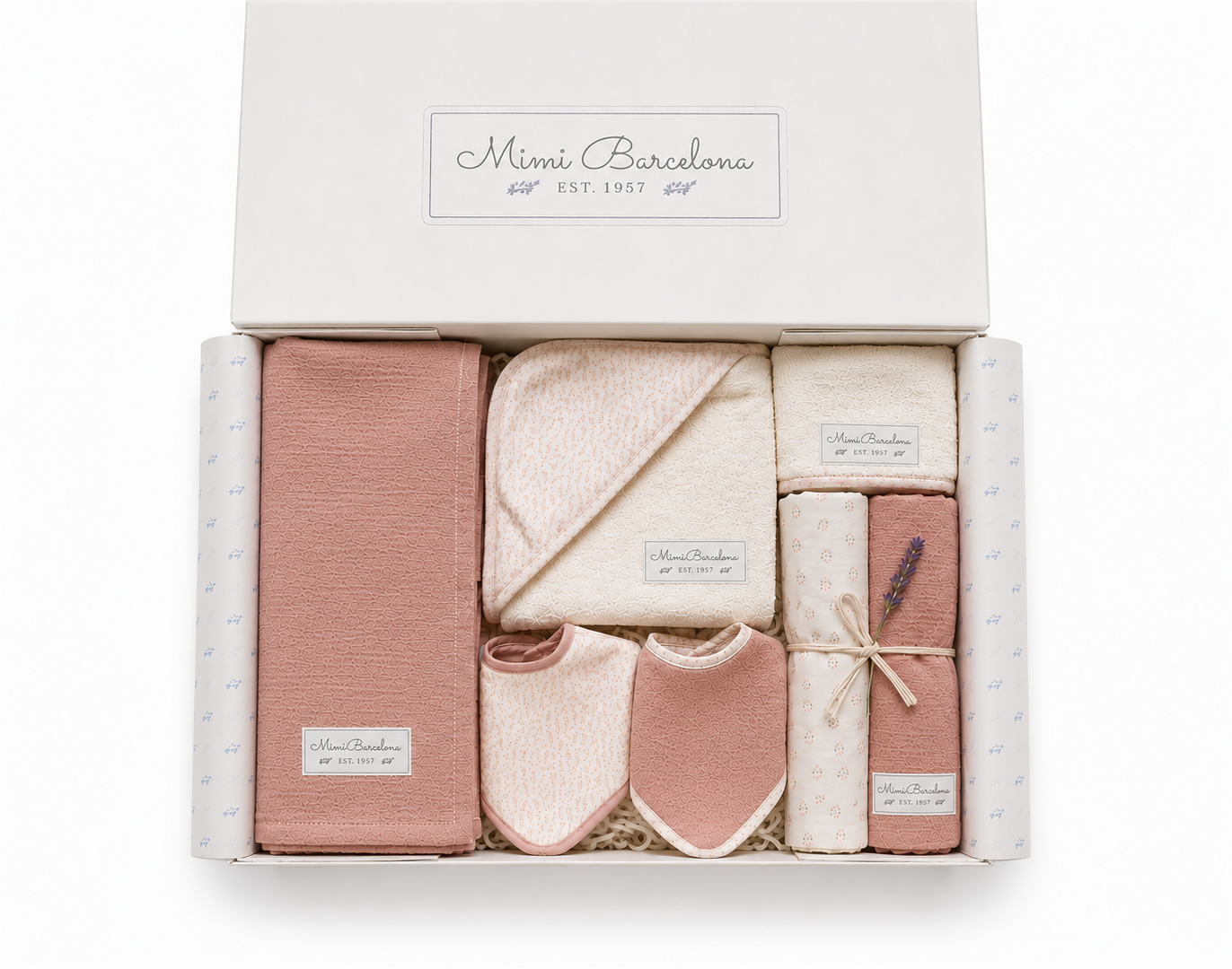 A baby gift set from Mini Barcelona in a white box, featuring a dusty pink blanket, a cream hooded towel, cream bibs, cream patterned socks, and a pink patterned wrap tied with a ribbon.