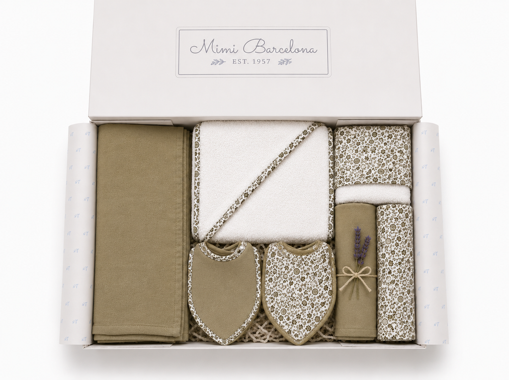 A baby gift set from Mimi Barcelona in a white box, featuring olive green and white patterned fabrics, including a blanket, a small bag with a chain strap, a tube with lavender, and two bibs.