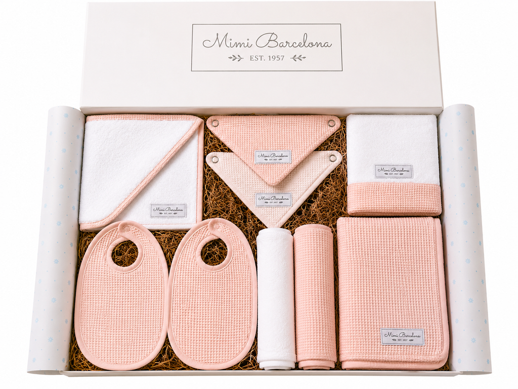 A baby gift set from Mimi Barcelona in a white box with brown shredded paper. The set includes pink and white textured baby cloths, including bibs, a blanket, a towel, and rolled fabric pieces.
