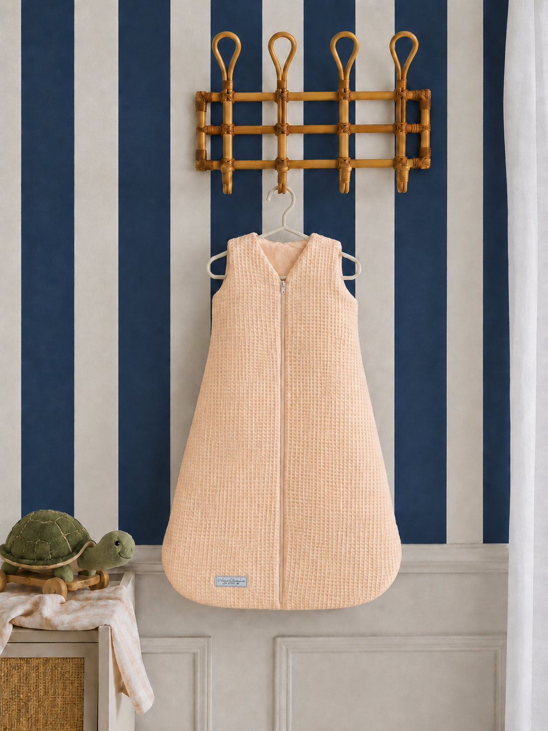 Beige baby sleeping bag hanging on a hook against a striped wall.