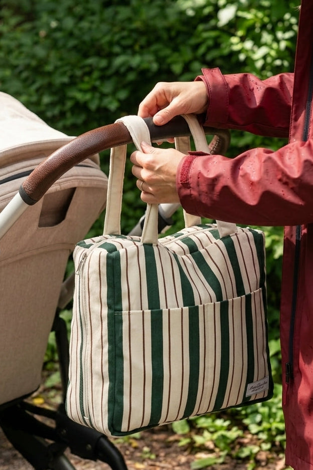 A person in a red jacket holds a green and cream striped diaper bag next to a stroller in a wooded outdoor setting.