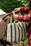 A person in a red jacket holds a green and cream striped diaper bag next to a stroller in a wooded outdoor setting.